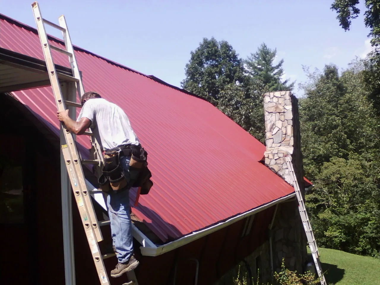 Roofer on a ladder beside a red metal roof and stone chimney — Soffit & Fascia Repair by Roofing Brothers in Shelter Island Heights